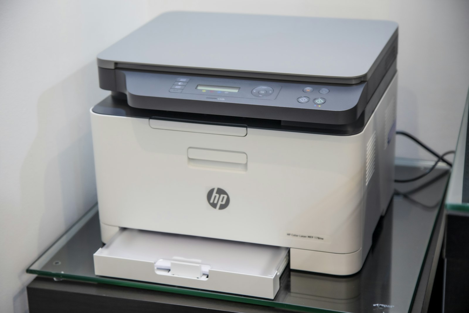 A white and grey all-in-one inkjet printer standing on a pale surface against a plain wall, lid closed, paper tray extended